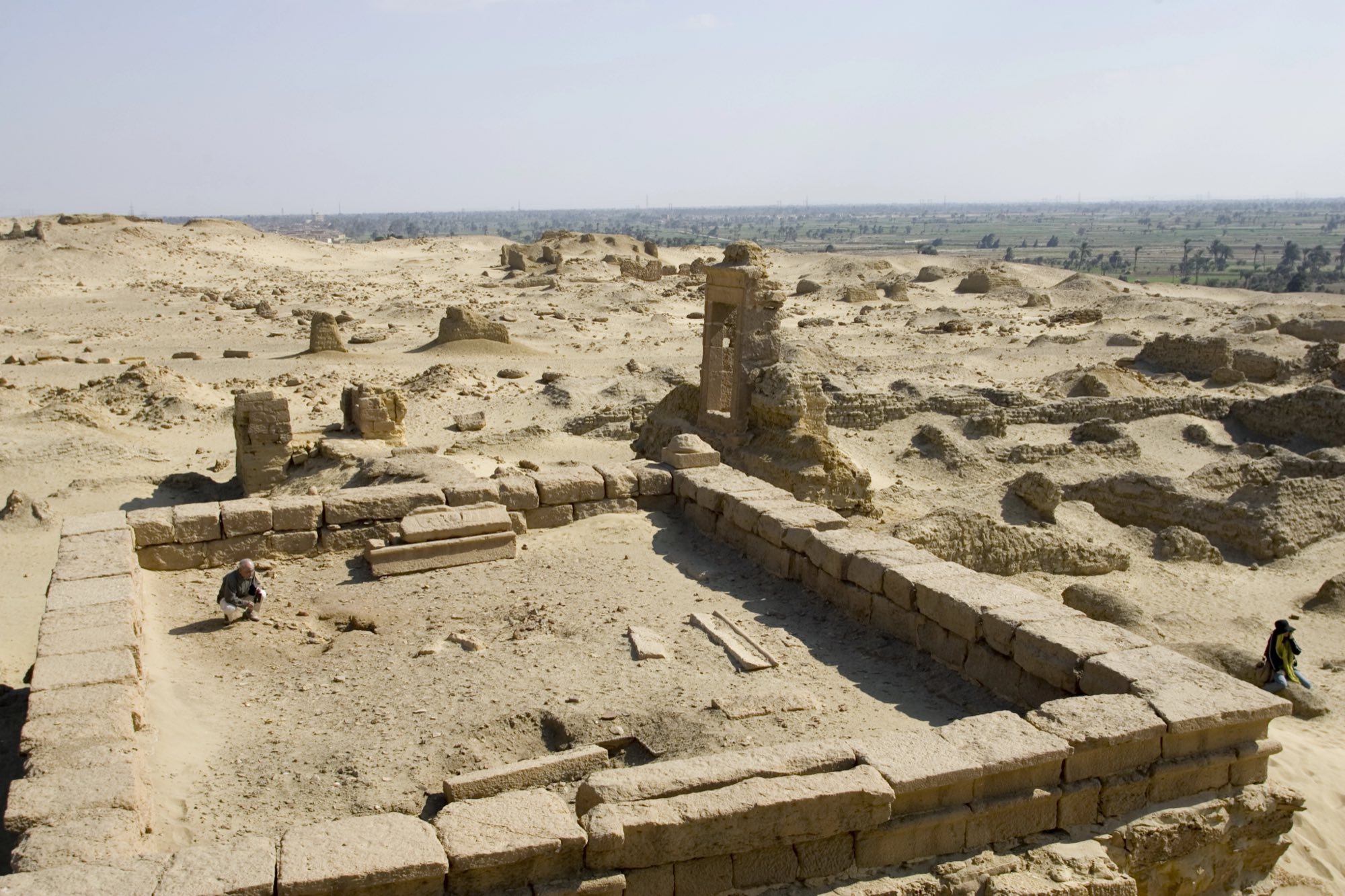 Ruined stone walls and a lone standing gateway at a Fayoum archaeological site, low desert mounds all around and the green edge of the oasis cultivation on the horizon